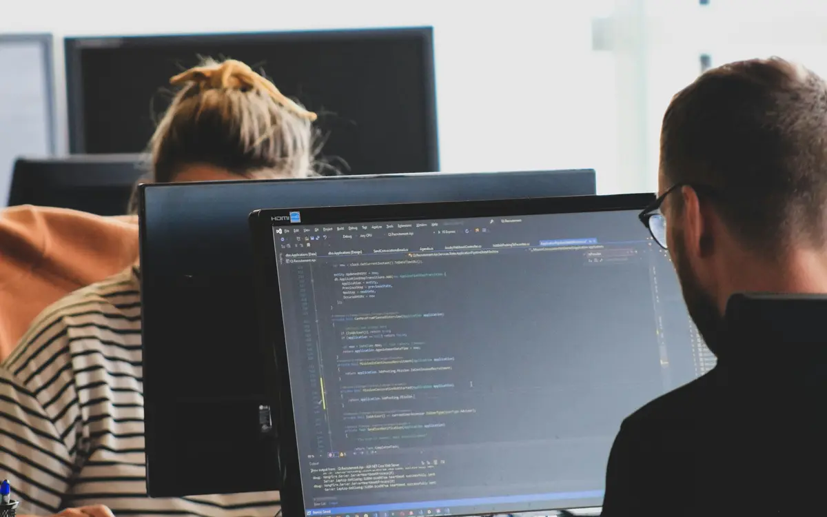 Duas pessoas programando lado a lado num escritório claro, com código na tela do monitor