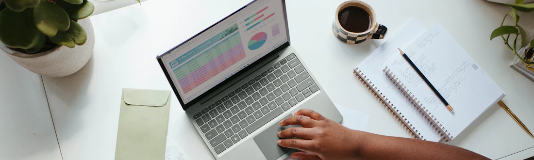 Notebook com uma planilha na tela, sobre uma mesa com caderno e café