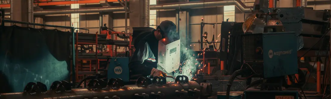 Soldador de máscara trabalhando numa peça de metal, com faíscas, no piso de uma fábrica
