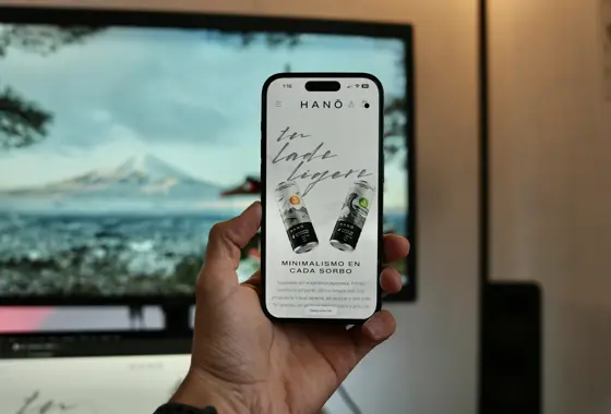 Mão segurando um celular com o site de uma marca de bebidas aberto, e um monitor desfocado ao fundo